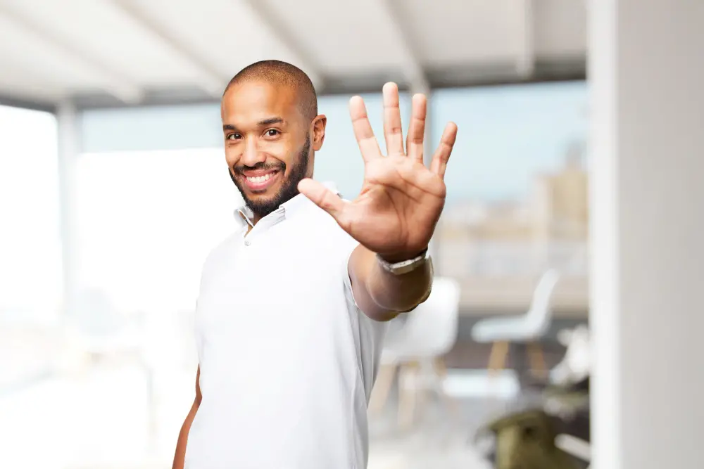 Homem sorrindo com a mão aberta em gesto de cumprimento em ambiente moderno.